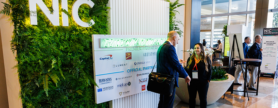 Two people at a conference stand in front of a sign highlighting sponsors of the event